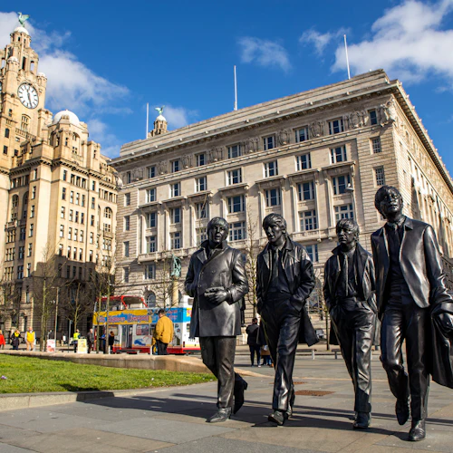 Bronsstatyer av fyra män som går, med historiska byggnader, grönt gräs och en dubbeldäckare i bakgrunden.