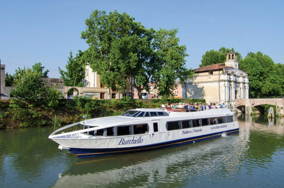 White passenger boat named "Burchiello" cruises on a canal with trees, buildings, and a clear sky in the background.