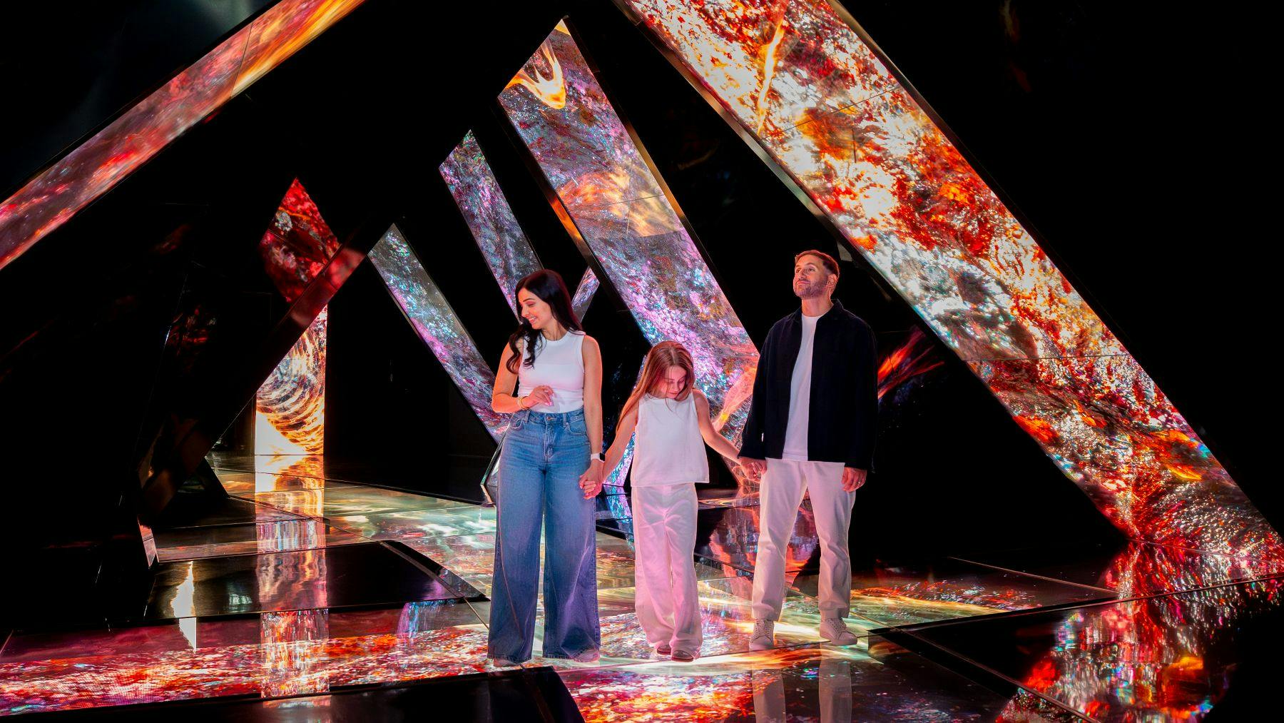 활기찬 추상적 패턴을 보여주는 각진 패널로 둘러싸인 반사 바닥에 세 사람이 서 있습니다.