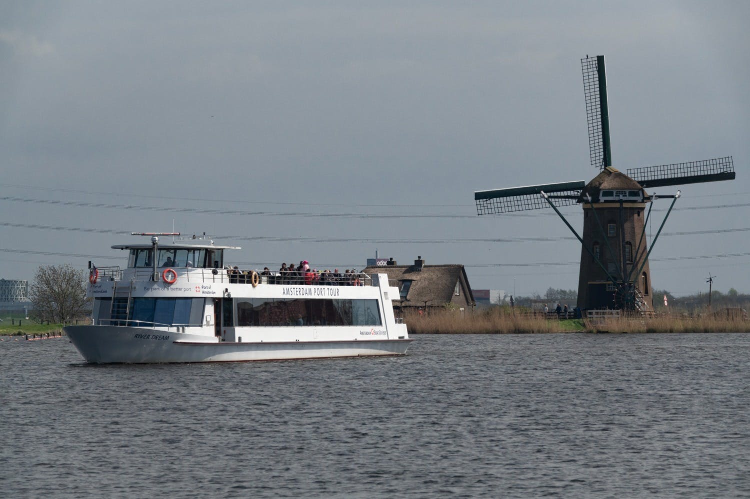 A river scene with a tour boat labeled “Amsterdam Port Tour” and a traditional windmill in the background near the shore.