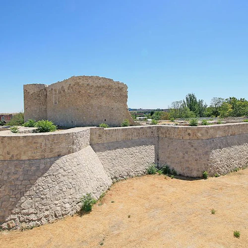 Forntida stenborg med höga murar, omgiven av ett torrt, sandigt område med gles vegetation och några närliggande byggnader.