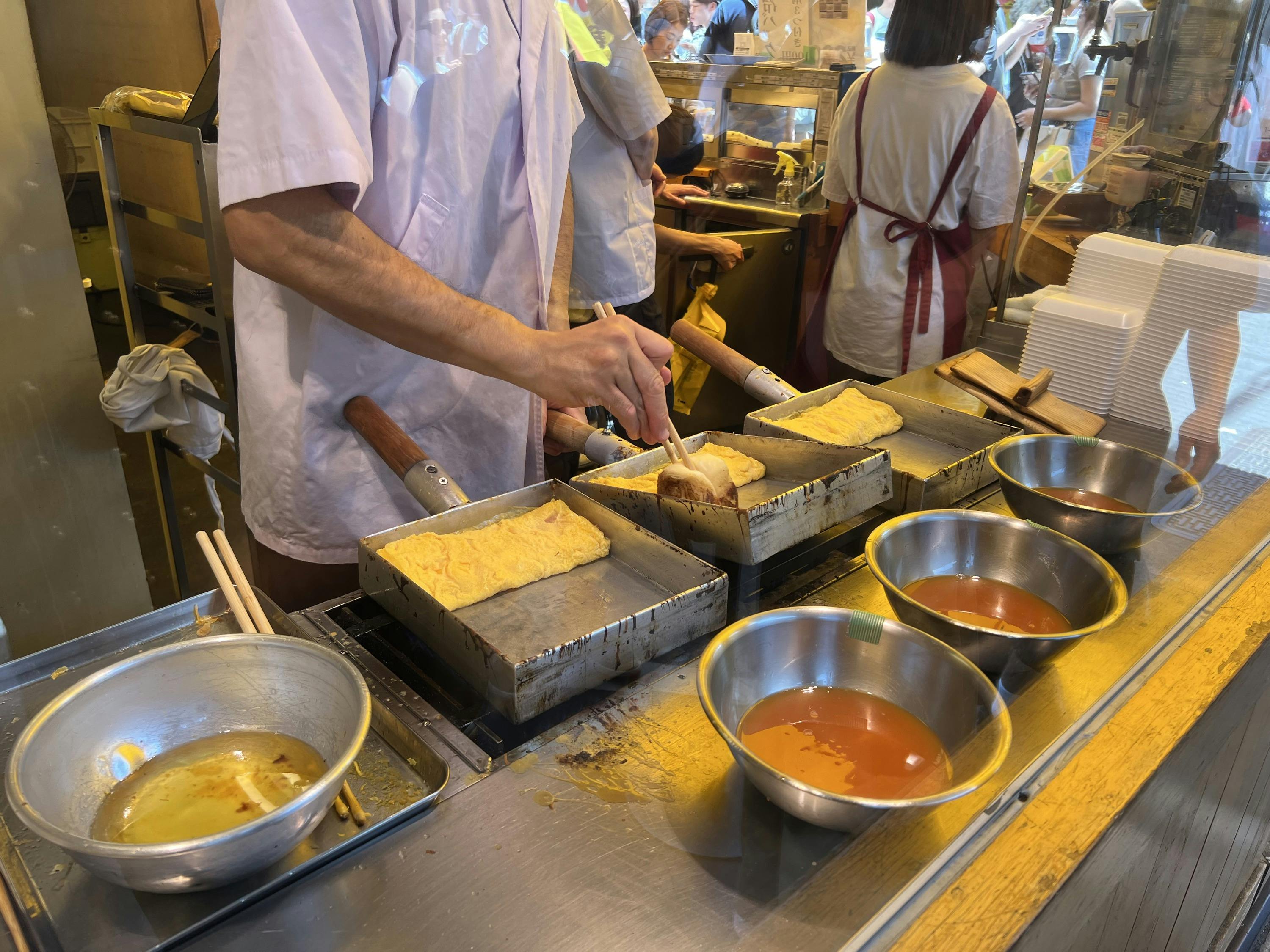 A person cooking tamagoyaki on rectangular pans at a food stall. Bowls with egg mixture and tools are visible on the counter.