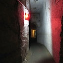 Dimly lit underground tunnel with rough walls, narrow path, and a red light fixture on the wall.
