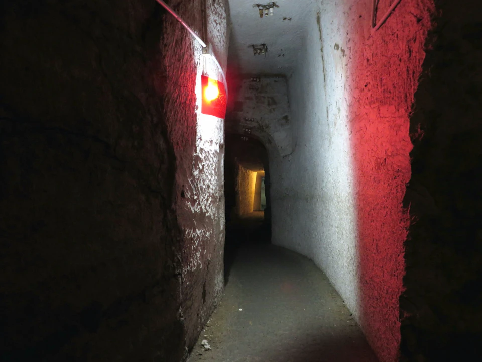 A dimly lit underground tunnel with rough walls, a narrow path and a red light fitting on the wall.