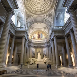 Stor sal med höga pelare, utsmyckat tak, stora statyer, väggmålningar och flera personer som går och observerar.