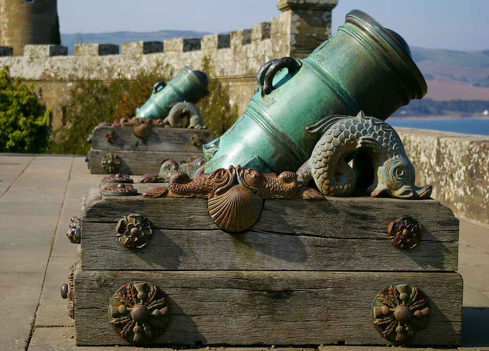Bronze cannons decorated with sea creatures on wooden platforms, set against a stone wall with scenic hills in the background.