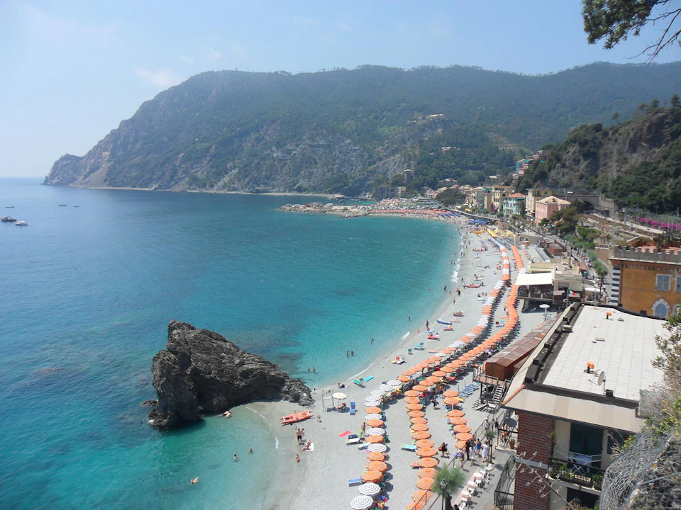 A coastal beach with rows of orange umbrellas and lounge chairs, clear blue water, and a rocky hill backdrop.