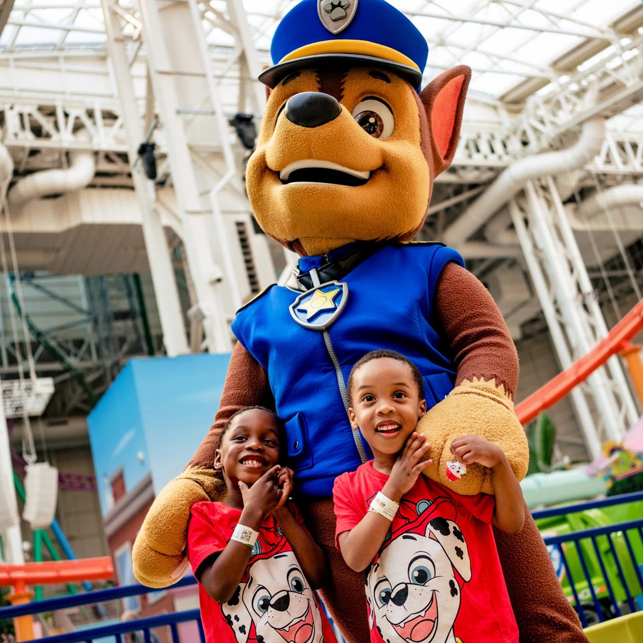 Two children smiling and posing with a person in a dog mascot costume wearing a police uniform in an indoor setting.