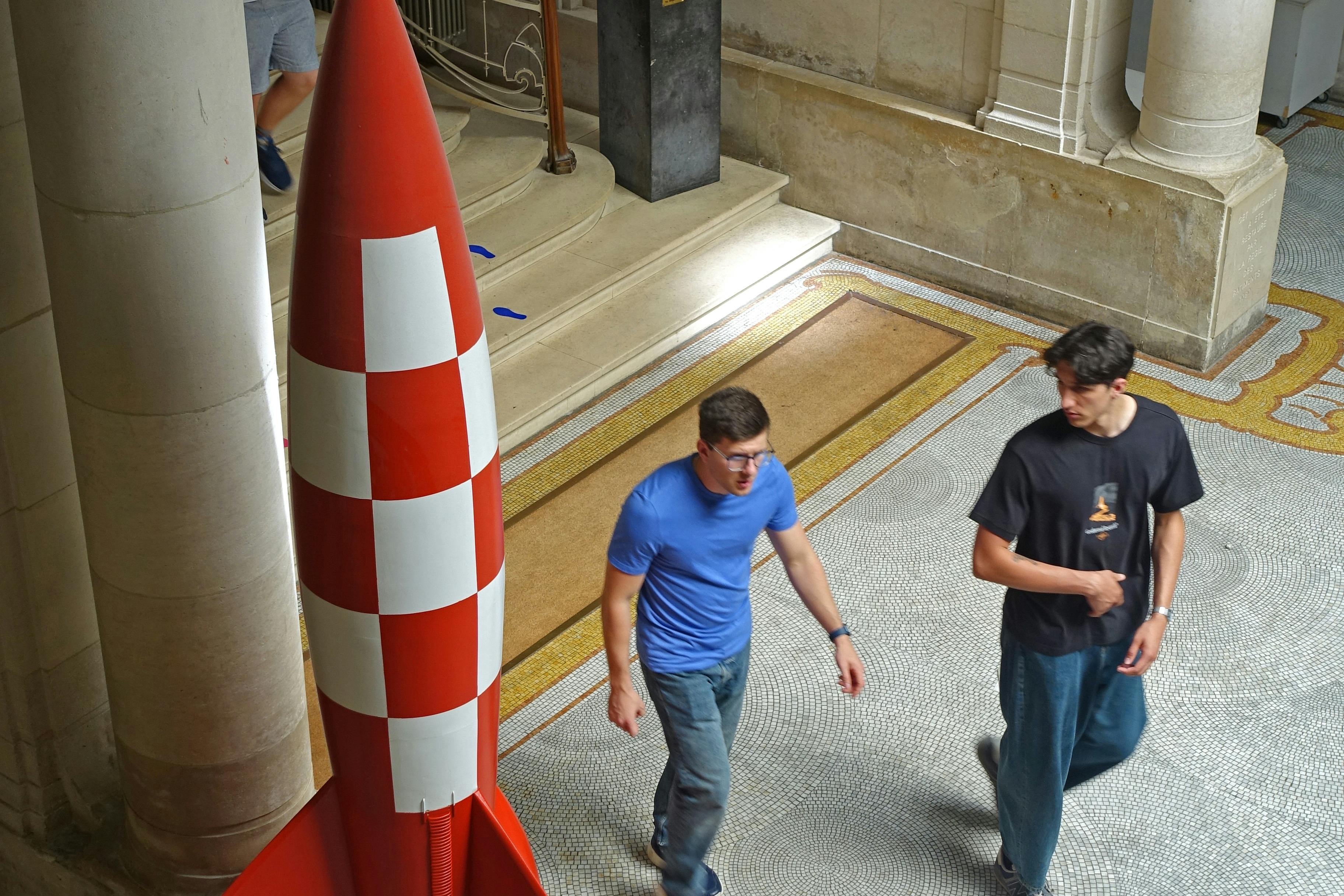 背の高い赤と白のロケット状の物体の近くのタイル張りの屋内空間を歩く2人の人物。