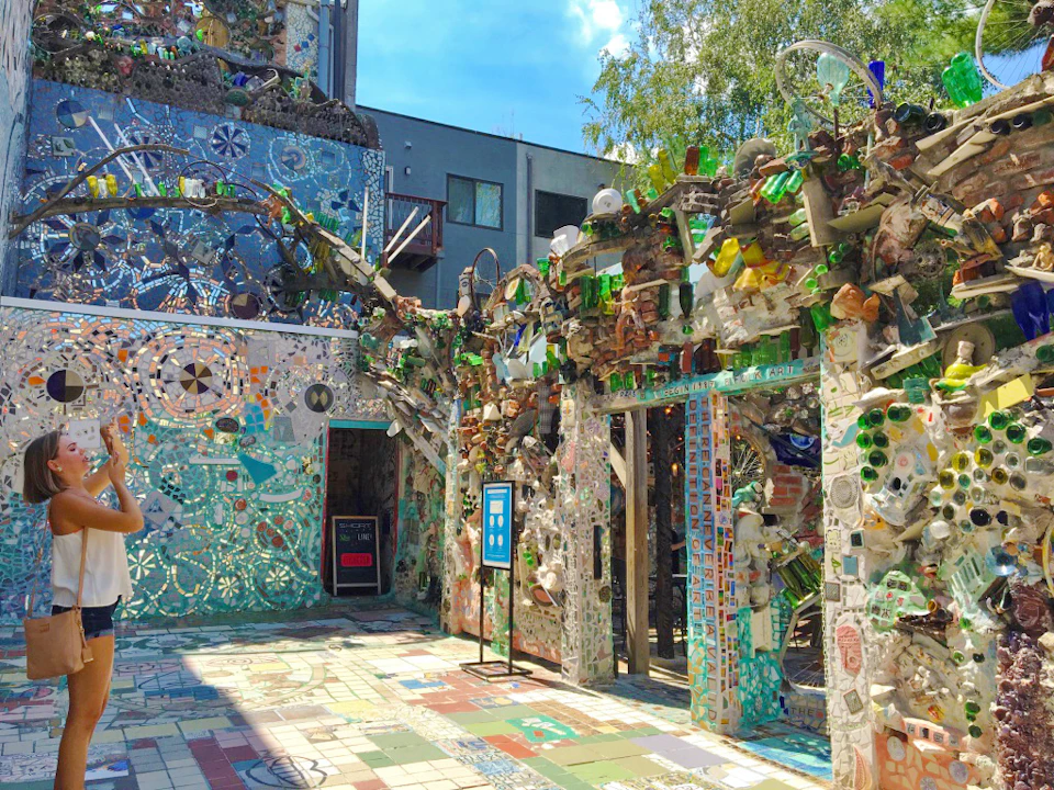 Colorful mosaic courtyard adorned with various objects and tiles, featuring a corridor entrance and an informational sign.