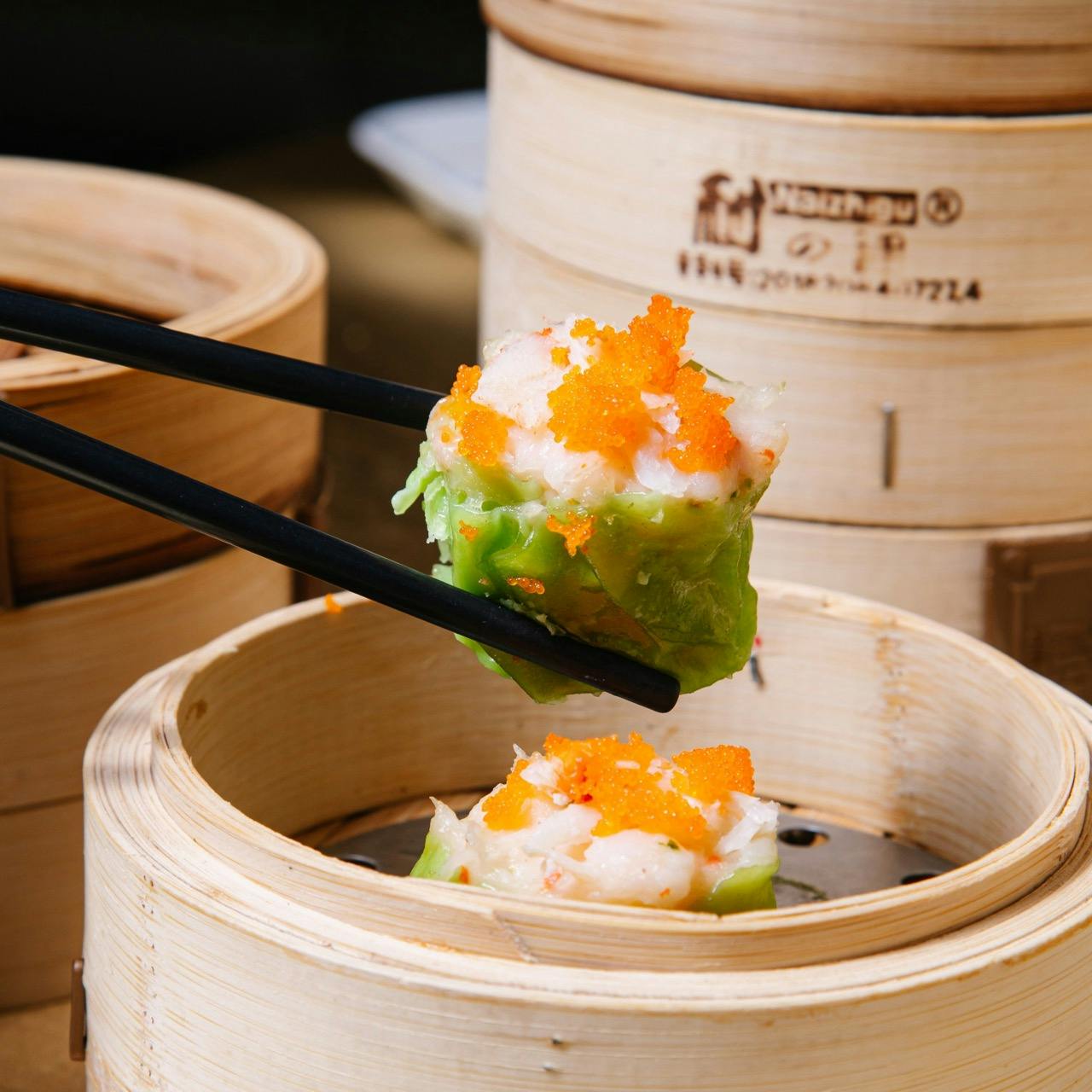 A pair of chopsticks holds a piece of dim sum topped with orange roe above a bamboo steamer with more dim sum.