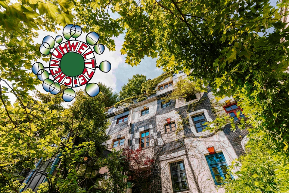 A multi-story building with colorful windows and ivy growing on its facade, surrounded by trees. A logo with the text "UMWELT ZEICHEN" is in the top left corner.