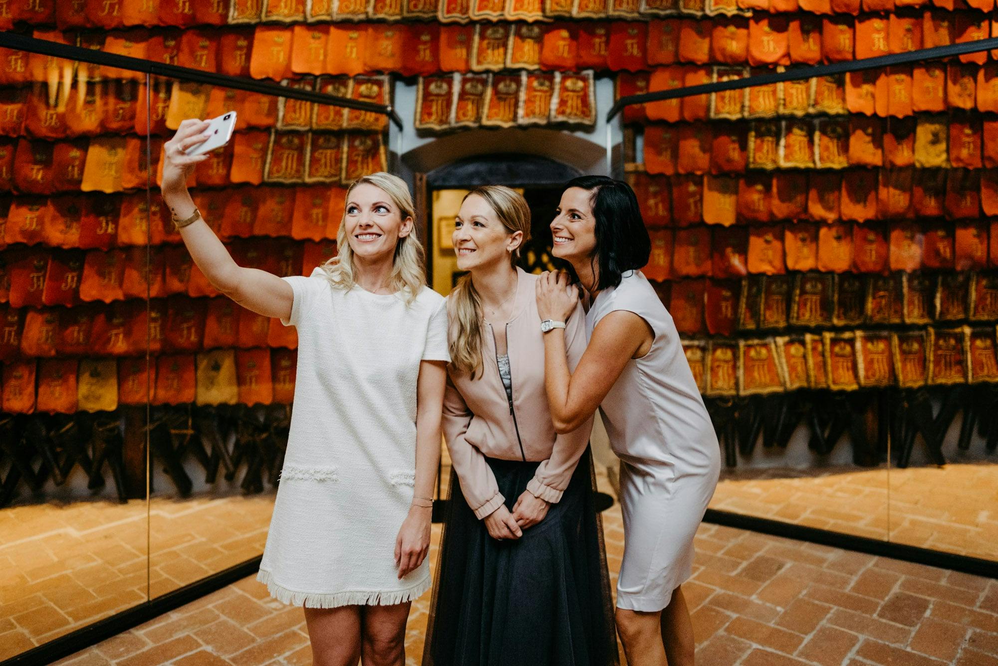 다채로운 색상의 전통 벽걸이로 장식된 방에서 세 명의 여성이 미소를 지으며 셀카를 찍으며 포즈를 취하고 있습니다.