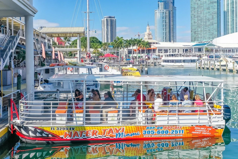 People seated on a water taxi in a marina, with tall buildings, palm trees, and other boats in the background.