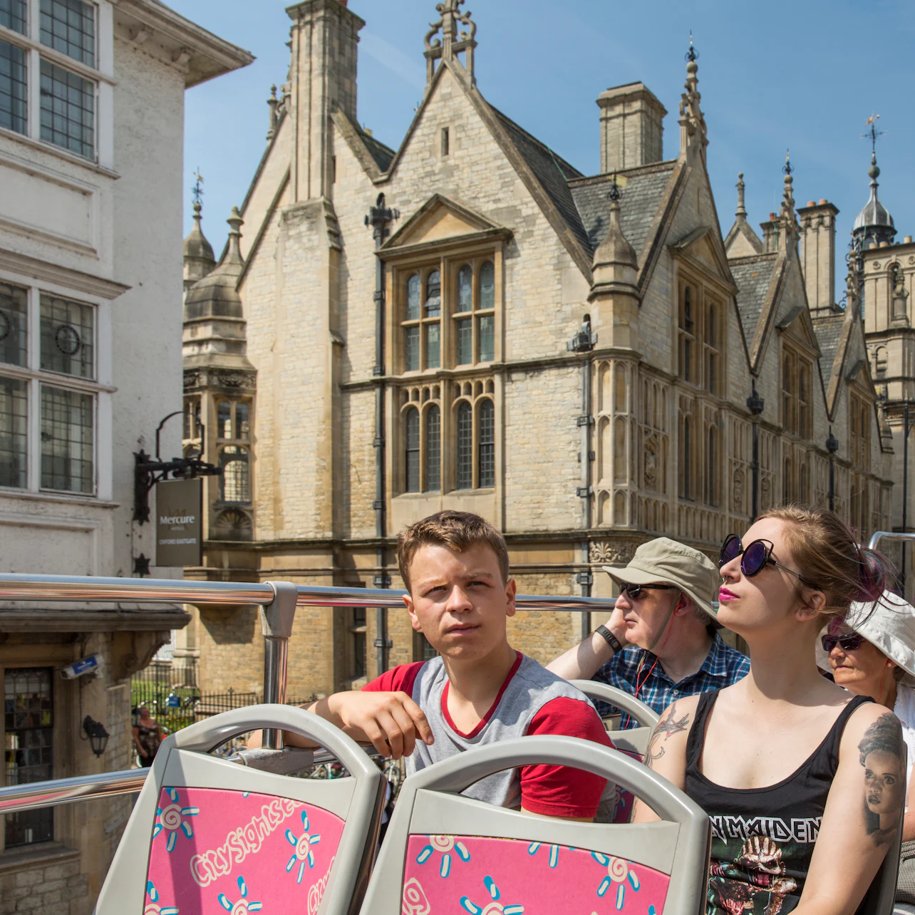 People sitting on the top deck of a tour bus, with historic stone buildings and a clear sky in the background.