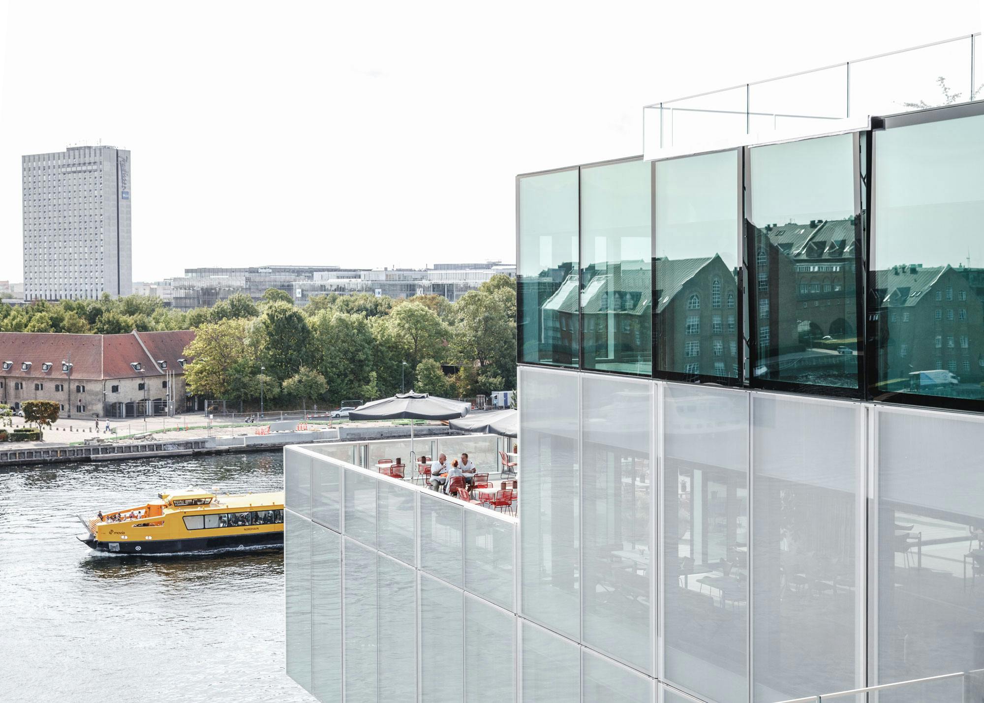 Un taxi aquàtic groc passa per un modern edifici de vidre amb terrassa amb vistes a l&#39;aigua. Al fons hi ha arbres i edificis.