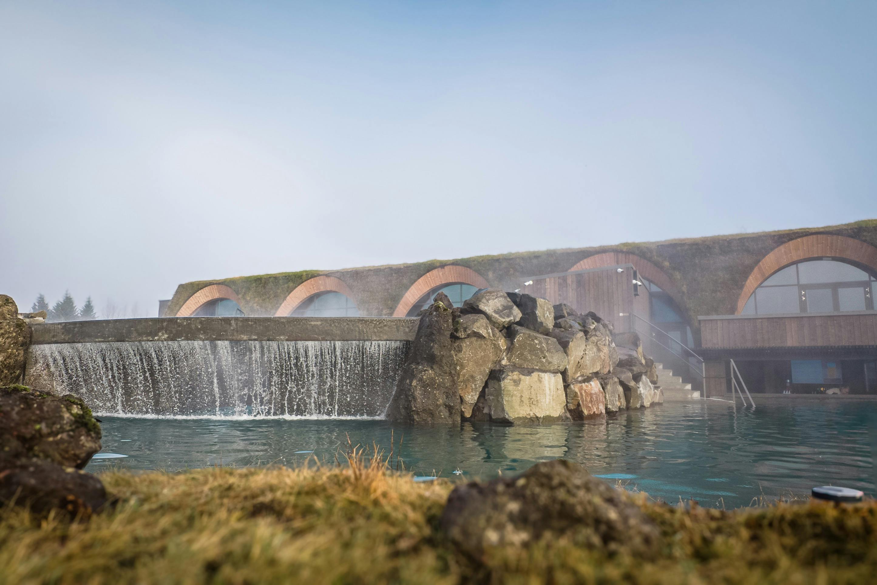 Een geothermische spa met een waterval en gewelfde gebouwen op de achtergrond, omgeven door rotsen en mist onder een heldere hemel.