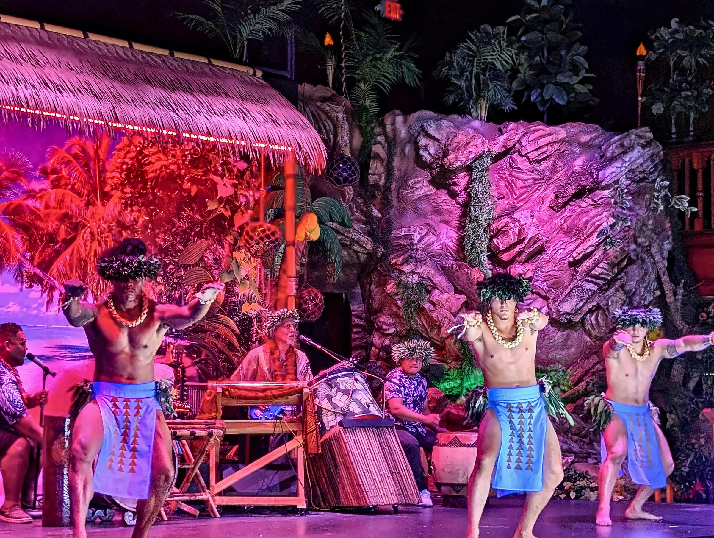 Performers in traditional Polynesian attire dance on a stage with tropical decorations and musicians in the background.