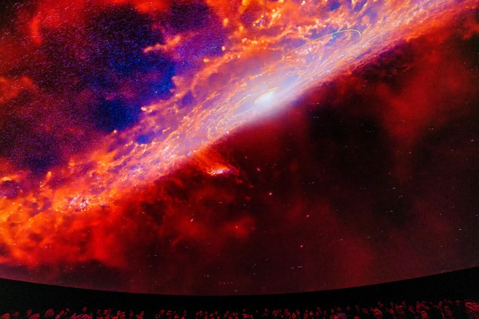 A vivid galaxy projection lights up a domed ceiling above a crowd, featuring swirling colors of red, blue, and white.