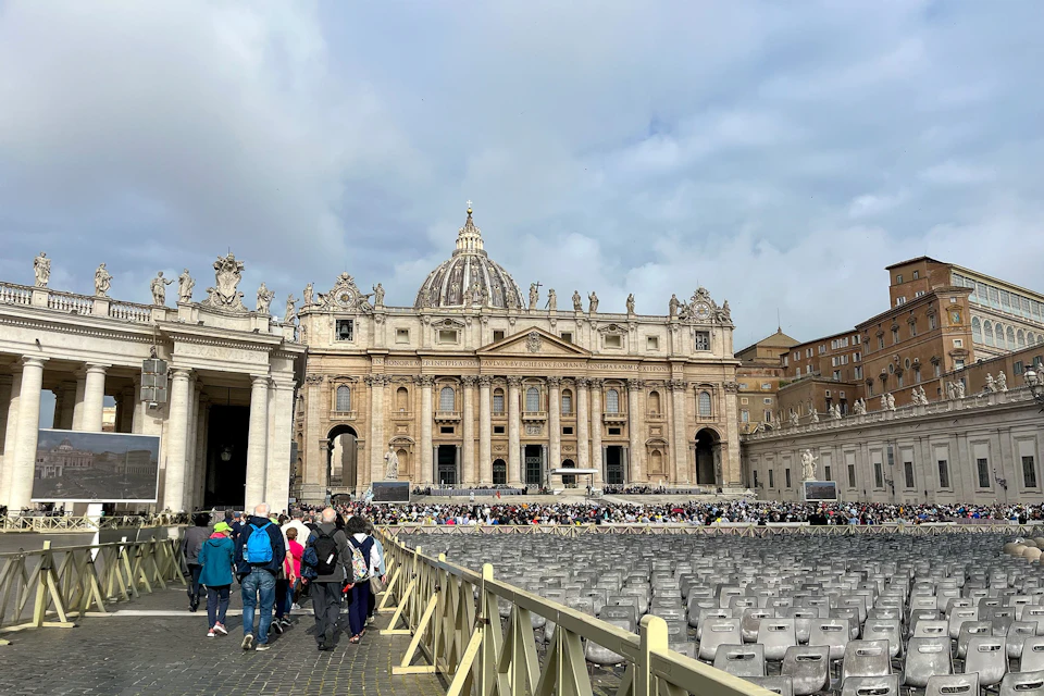 A view of St. Peter's Basilica in Vatican City with a crowd of people and rows of chairs in the foreground.