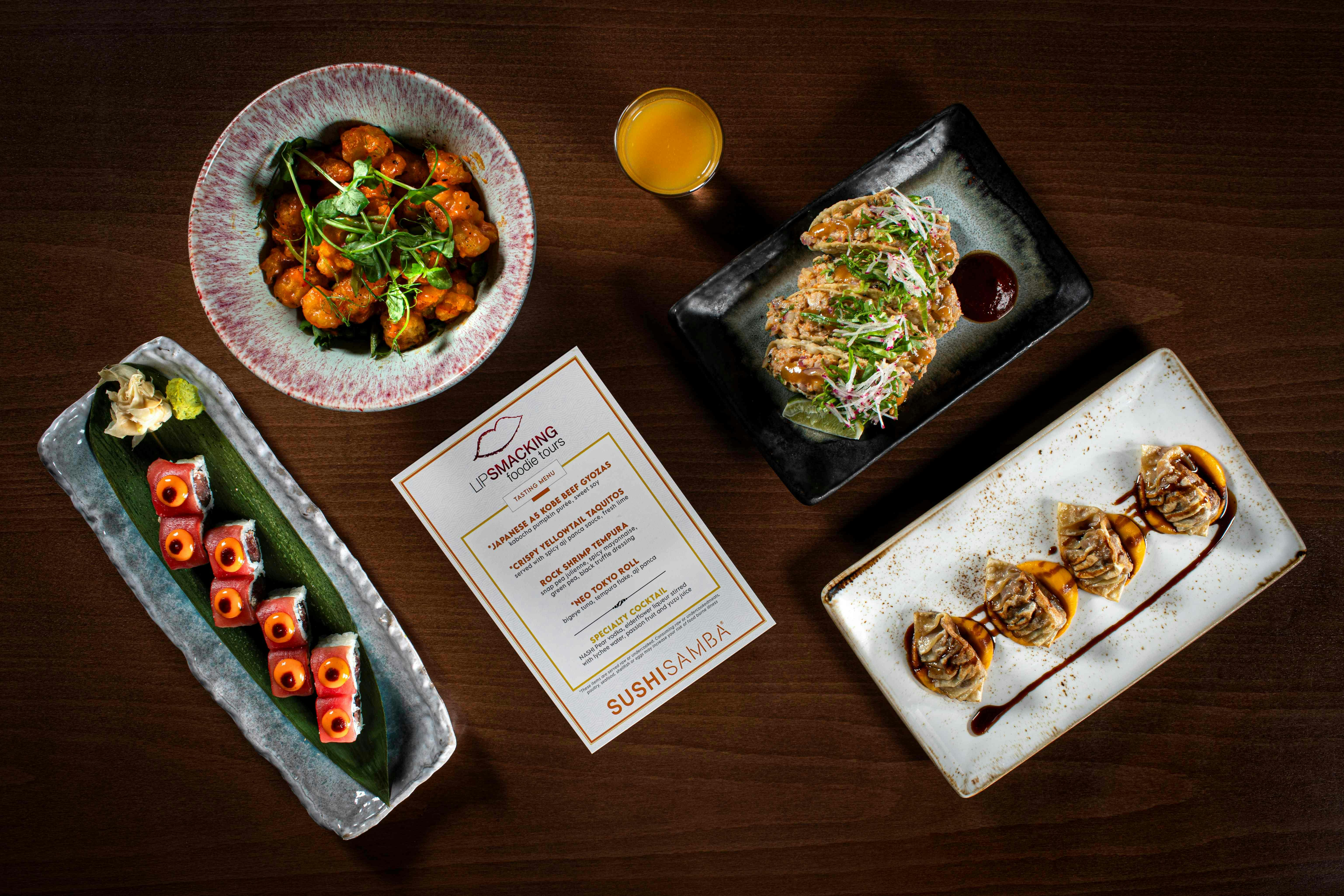 Ένα τραπέζι με διάφορα πιάτα, ένα ποτό και μια κάρτα μενού από το Sushisamba.