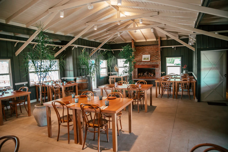 A cozy restaurant interior with wooden tables and chairs, potted plants, and a brick fireplace under a vaulted ceiling.