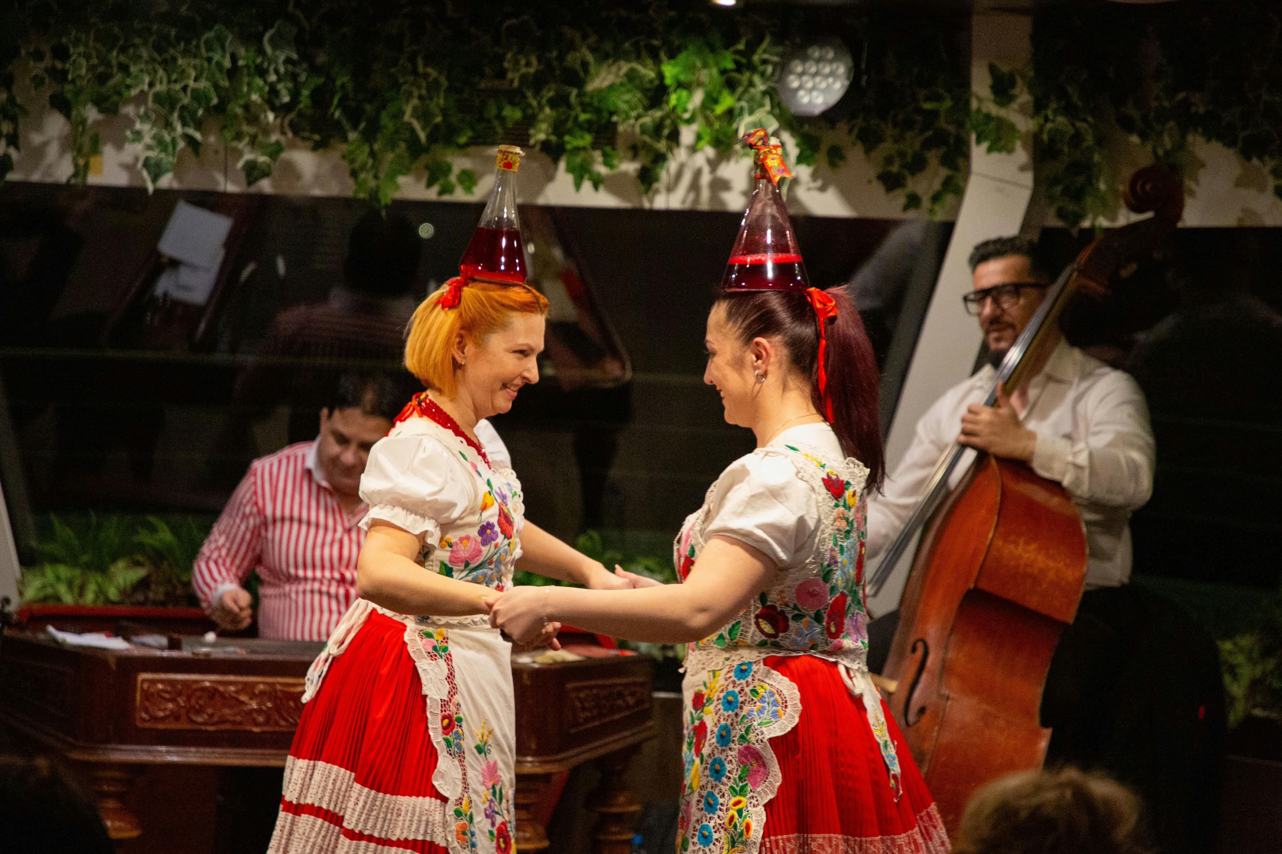 Two women in traditional dresses and unique hats dance, while a musician in the background plays an instrument.