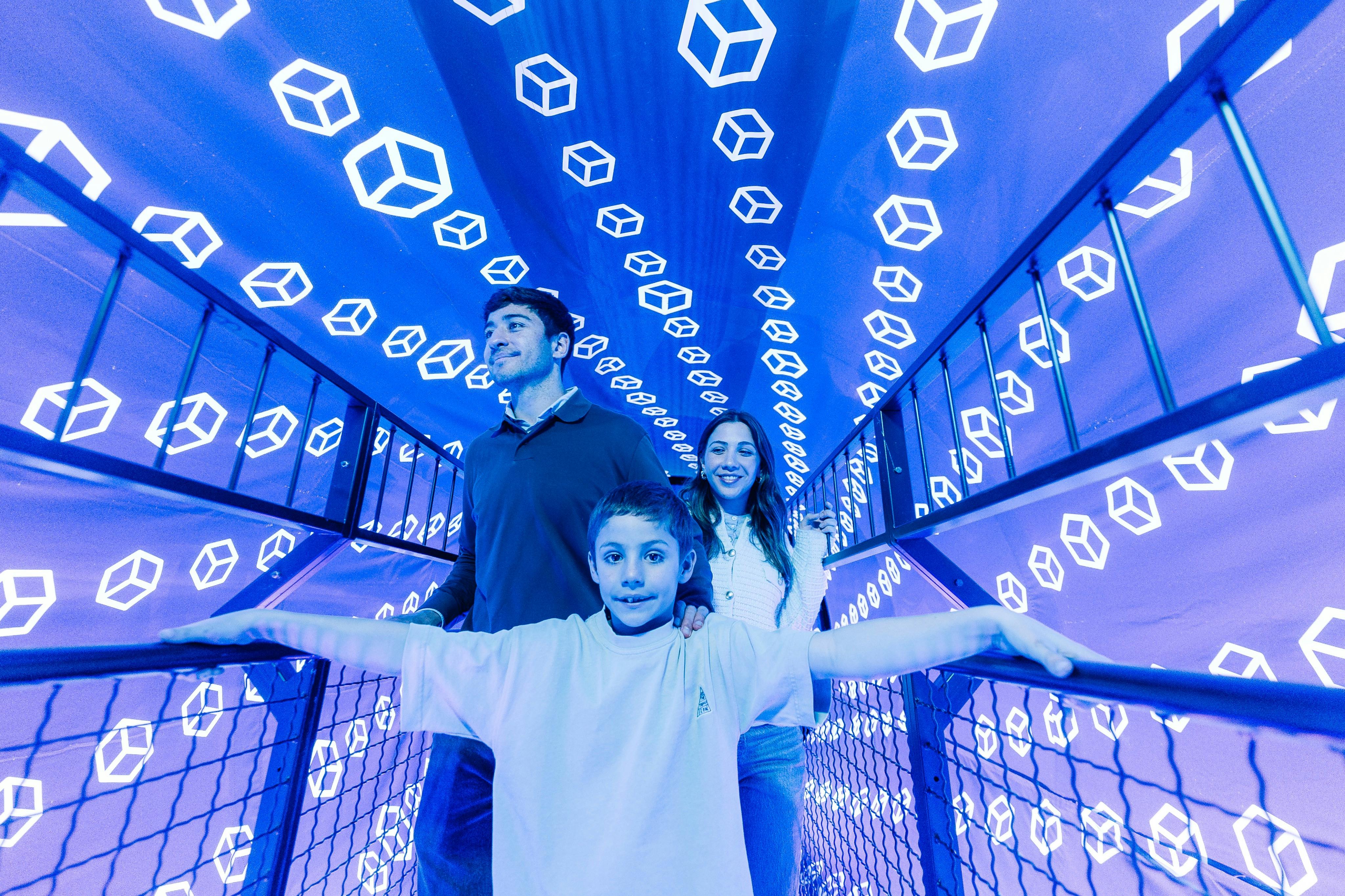 Chlapec s nataženýma rukama stojí před usmívajícím se mužem a ženou uvnitř tunelu s plovoucími geometrickými tvary na modrých stěnách.