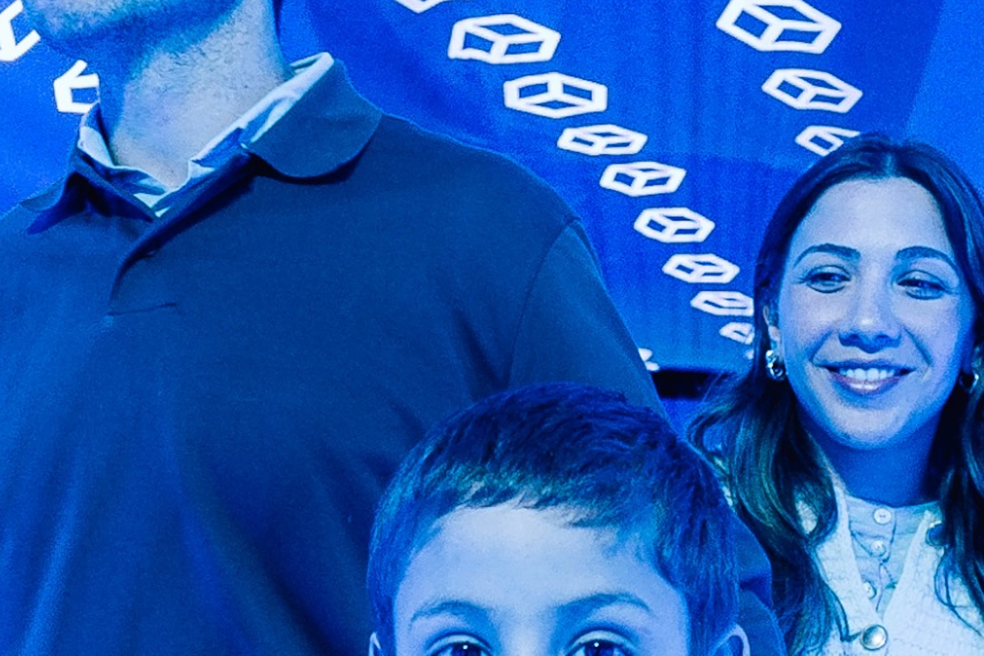 A boy with outstretched arms stands in front of a smiling man and woman inside a tunnel with floating geometric shapes on blue walls.