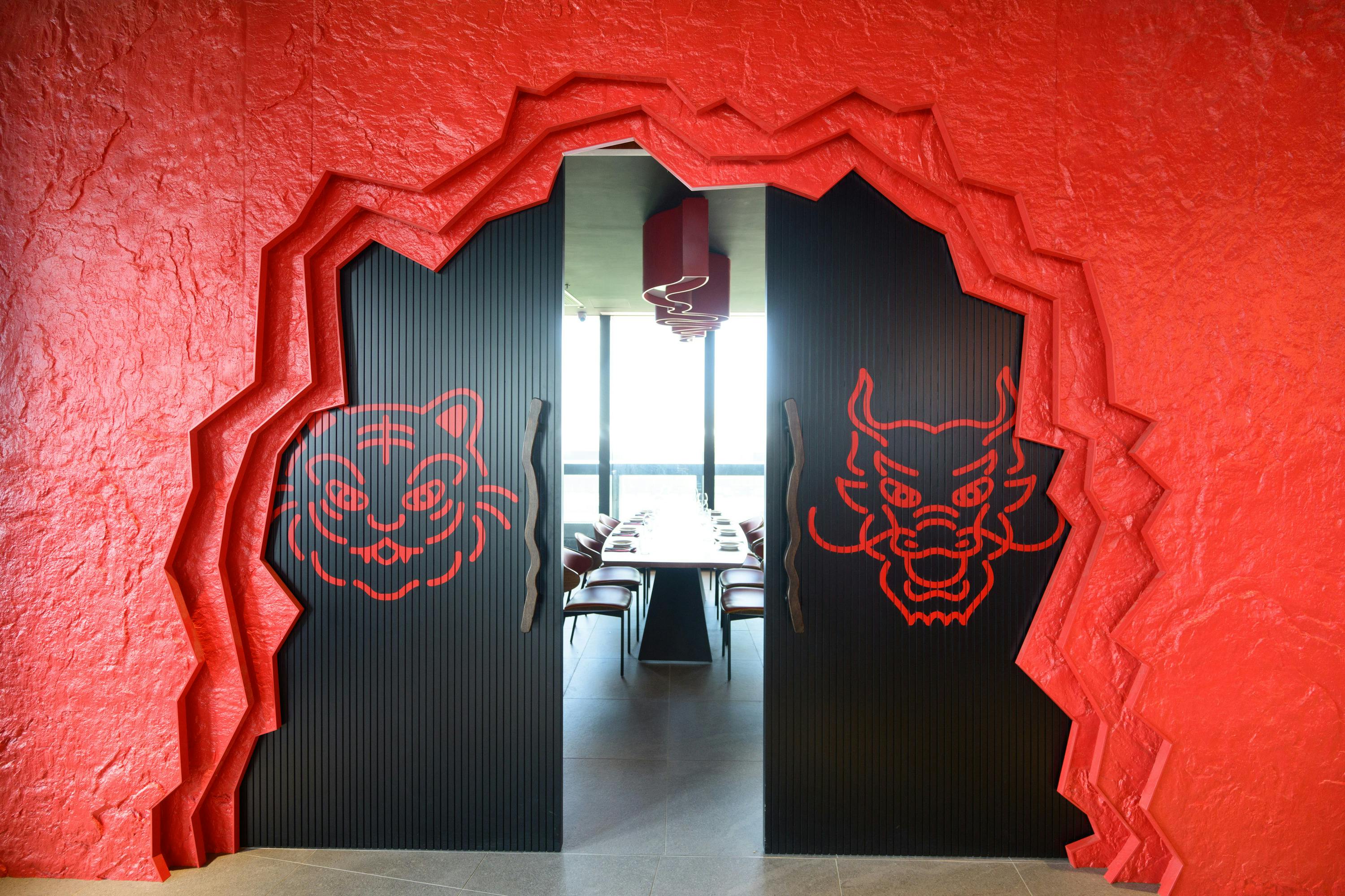 A restaurant entrance with red walls featuring tiger and dragon motifs on black doors, leading to a dining area with a long table.