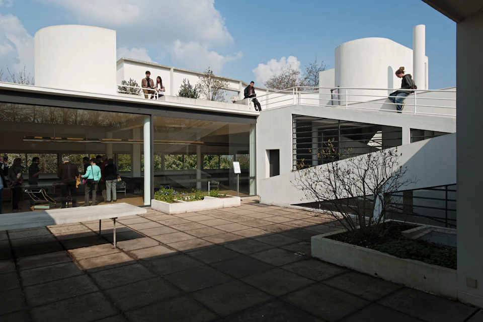 Modern white building with large windows, rooftop terrace with people, and open courtyard with plants. Clear blue sky above.