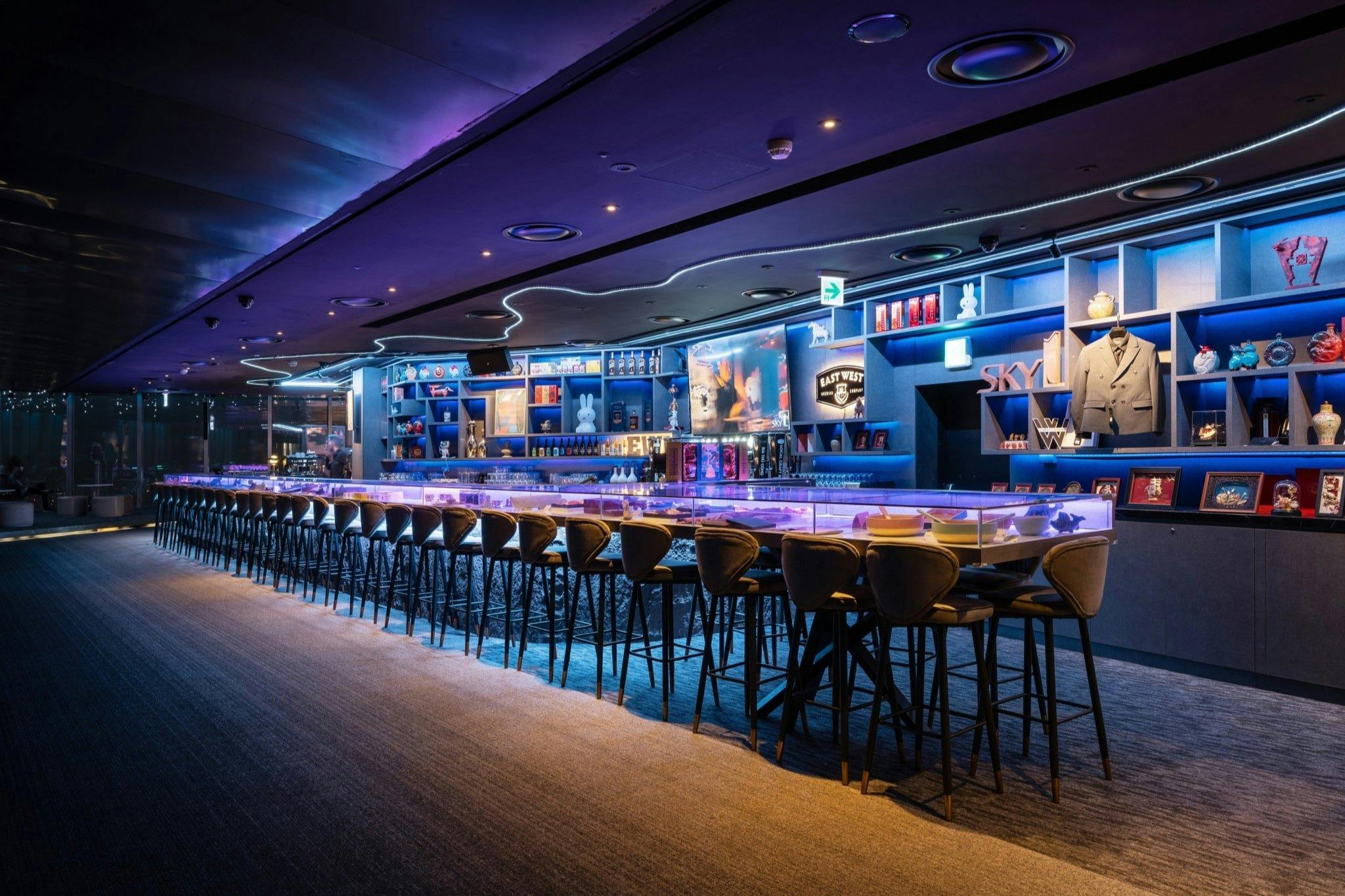 A sleek, modern bar with illuminated counters, high stools, and shelves stocked with bottles and glassware under glowing blue lights.