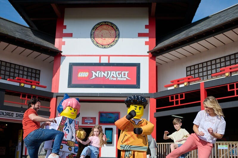 Uma personagem LEGO Ninjago e uma família a posar em frente à atração Ninjago The Ride num parque temático.