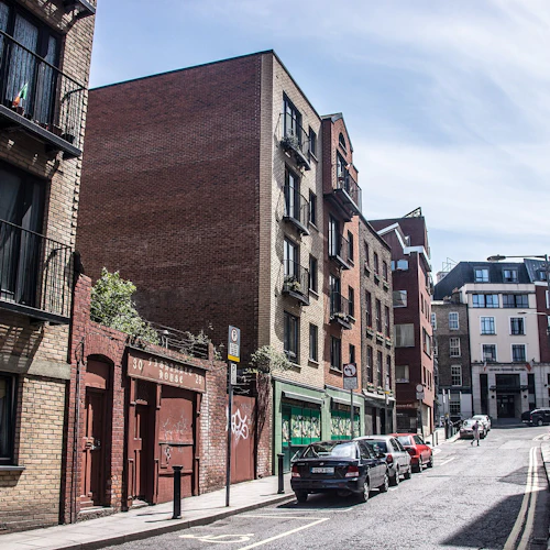 Smal gata med parkerade bilar och flerbostadshus på båda sidor, vilket leder till en avlägsen modern struktur under en molnig himmel.