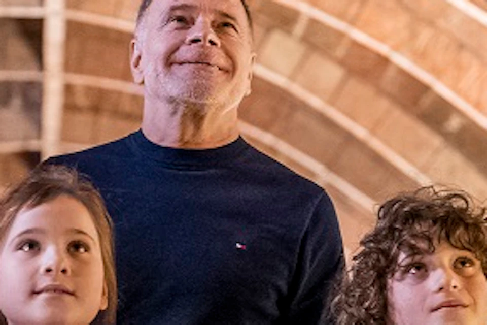 A family of four, consisting of parents and two children, stands under an arched ceiling, holding hands and looking upwards.