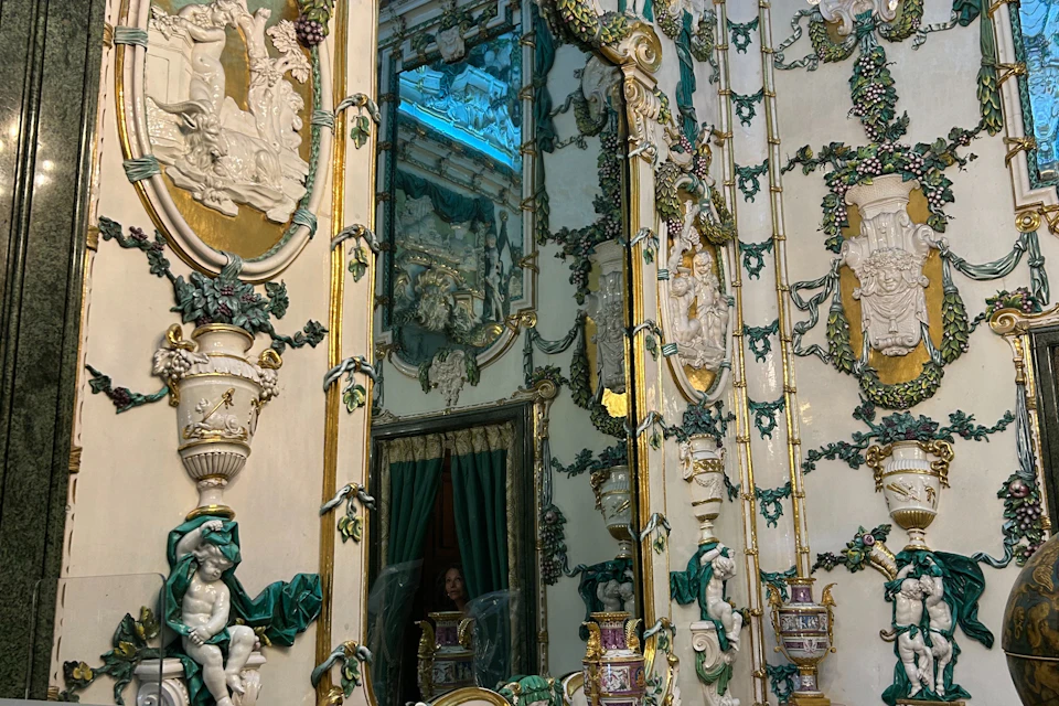 An ornate interior with intricate decorations, mirrors, and green drapery. A person is partially visible in the background.