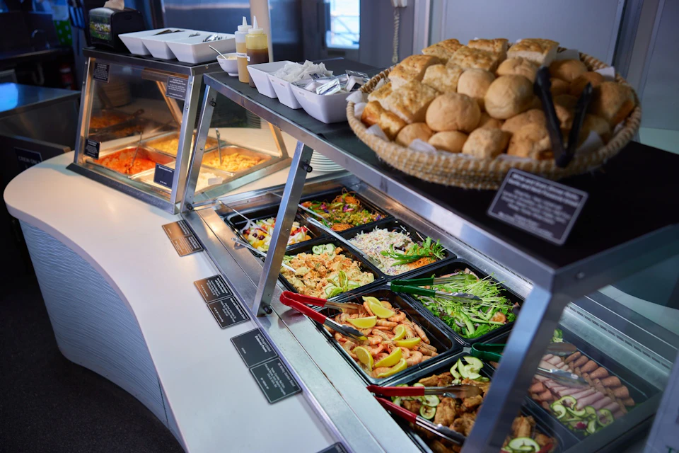 Salad bar with various fresh vegetables, fruits, and condiments alongside a basket of bread rolls and dispensers for sauces.