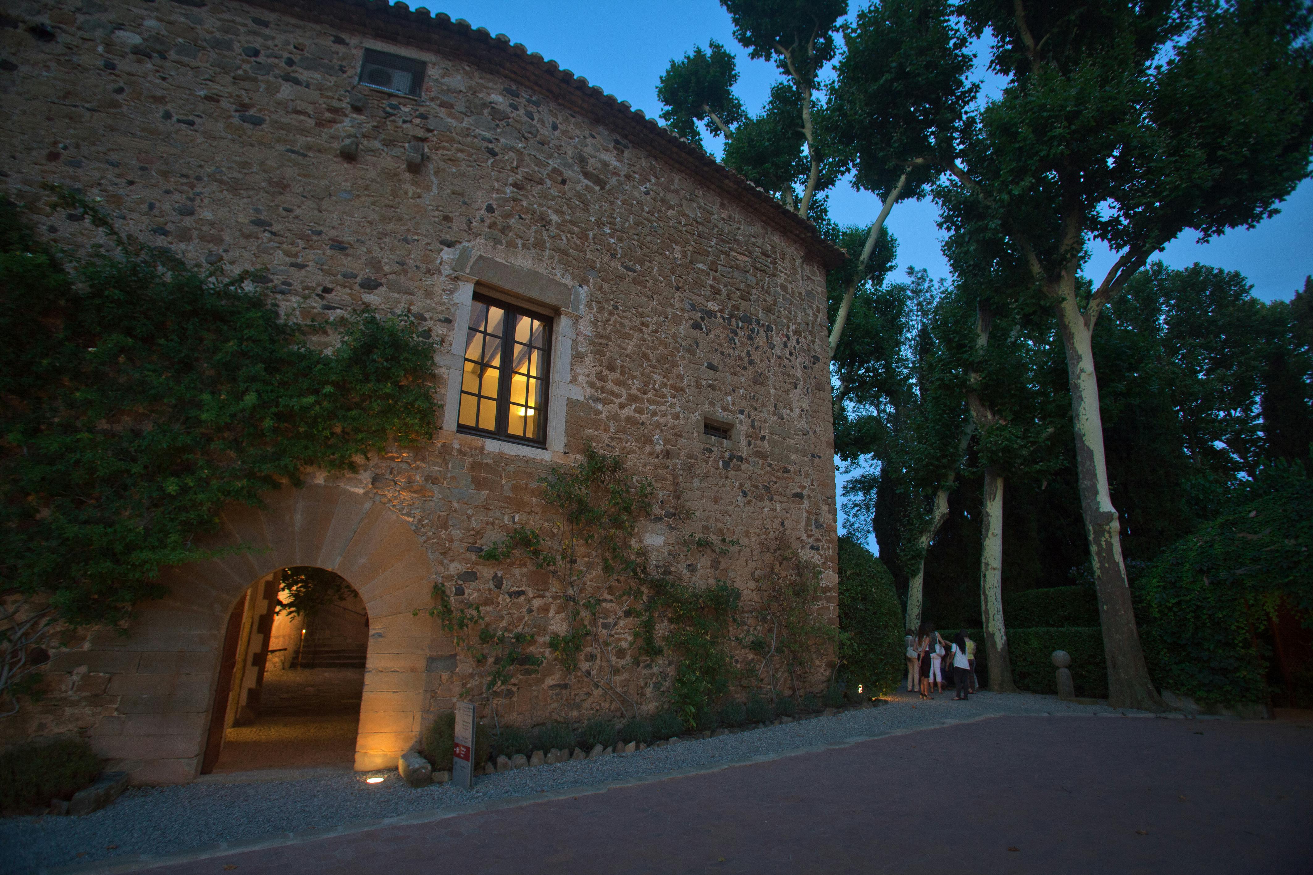 Каменное здание с освещенным окном и арочным дверным проемом. Плющ растет на стенах. Группа людей, стоящих снаружи. Деревья окружают сцену.