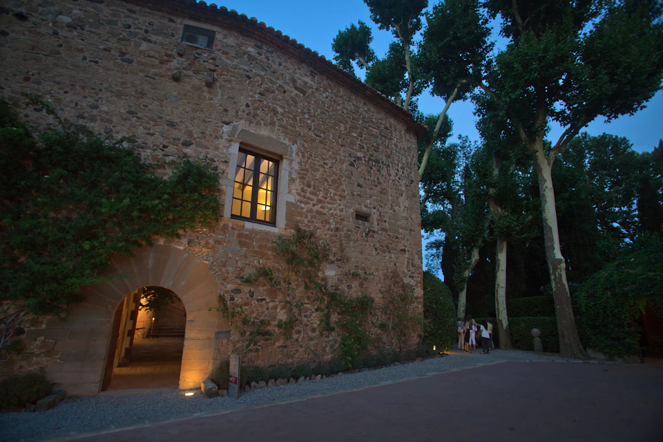 Stone building with a lit window and arched doorway. Ivy grows on walls. Group of people standing outside. Trees surround the scene.
