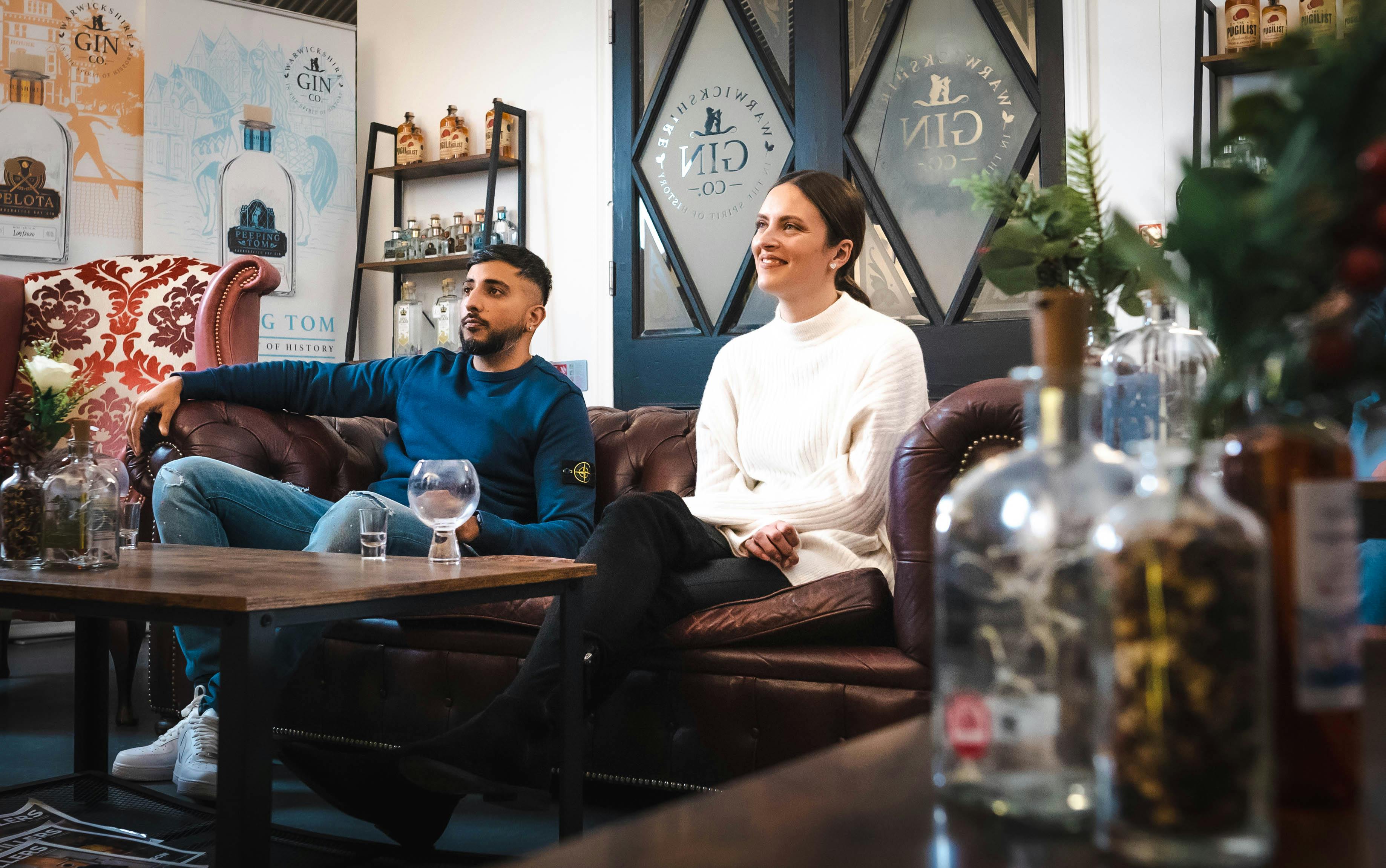 Dva lidé sedící na kožené pohovce v lihovarnickém salonku s lahvemi ginu na stole a policích a značkovými plakáty za sebou.