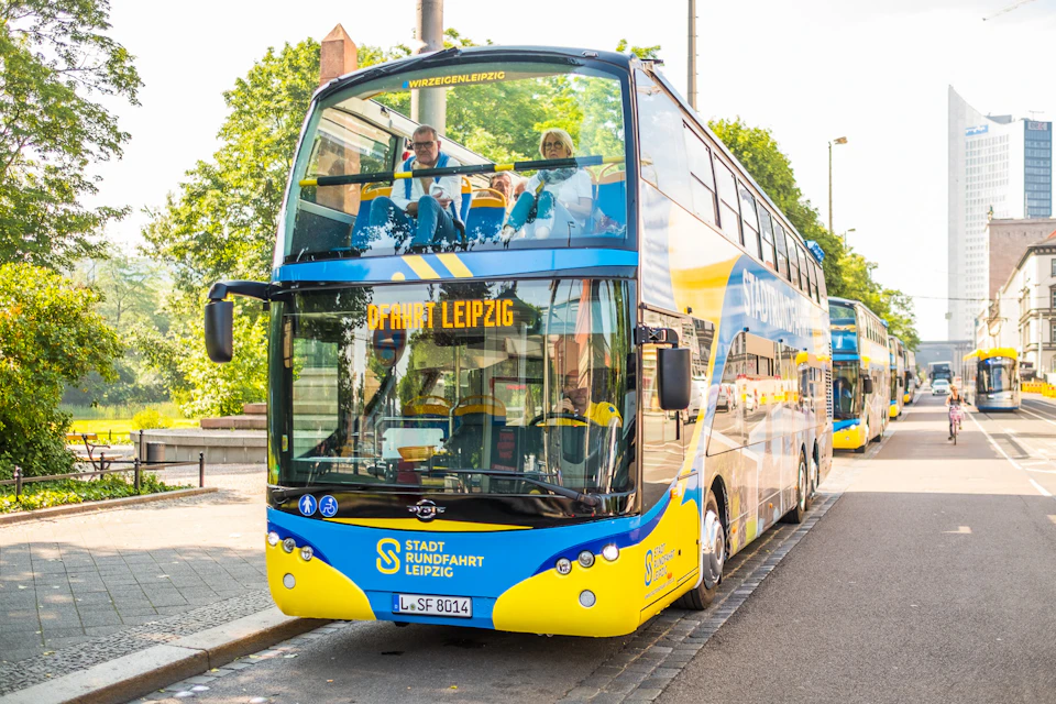 Double-decker tour bus in Leipzig with passengers on the upper deck, parked at the curb on a sunny day.