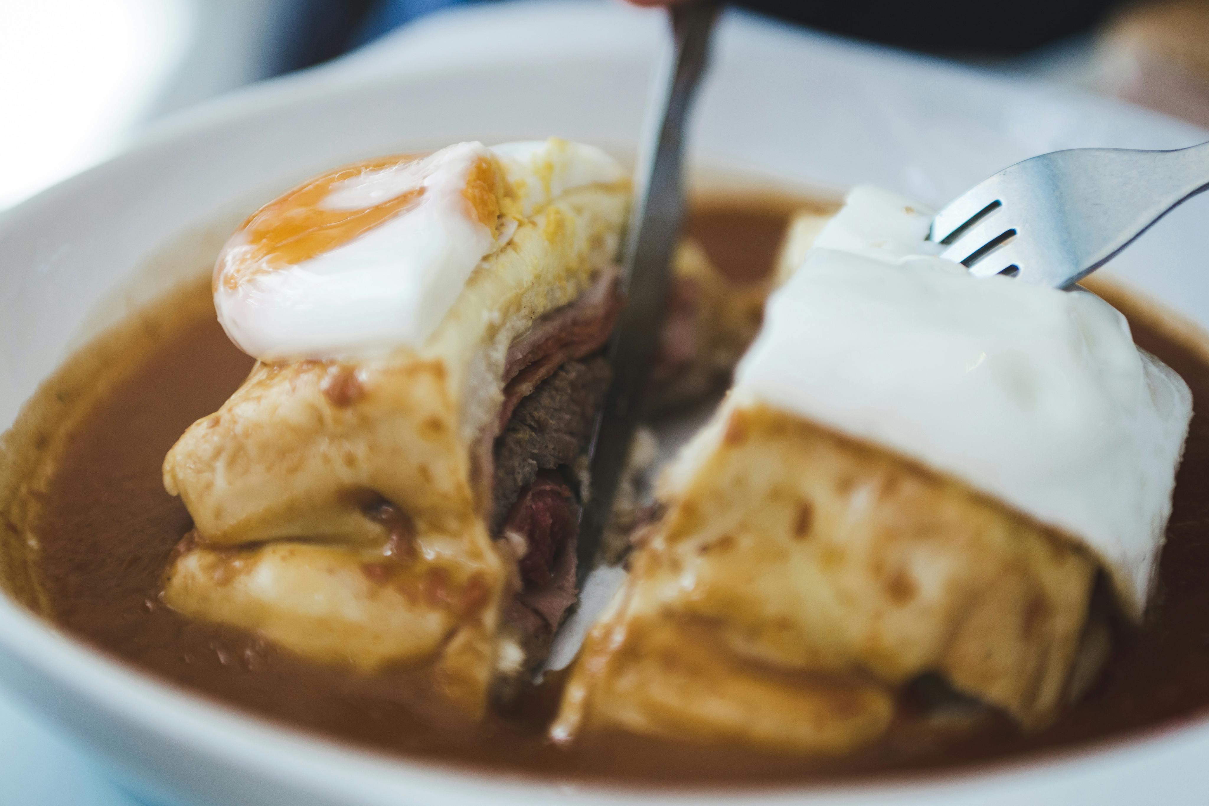 Close-up van een persoon die in een sandwich snijdt met laagjes vlees, kaas, een ei en saus op een wit bord.