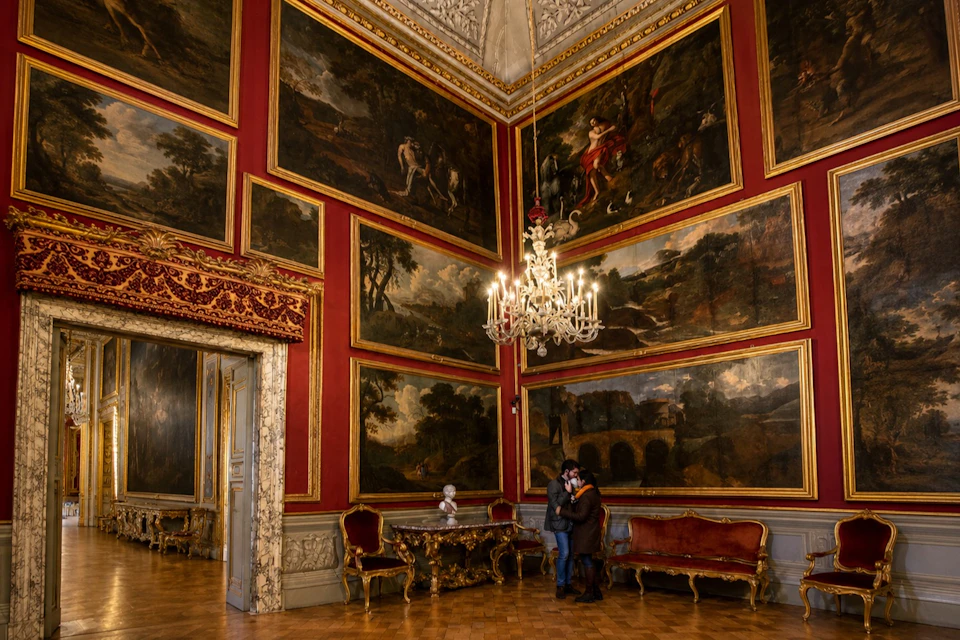 An ornate room with red walls, large framed paintings, and a chandelier. A couple stands near a red sofa and wooden furniture.