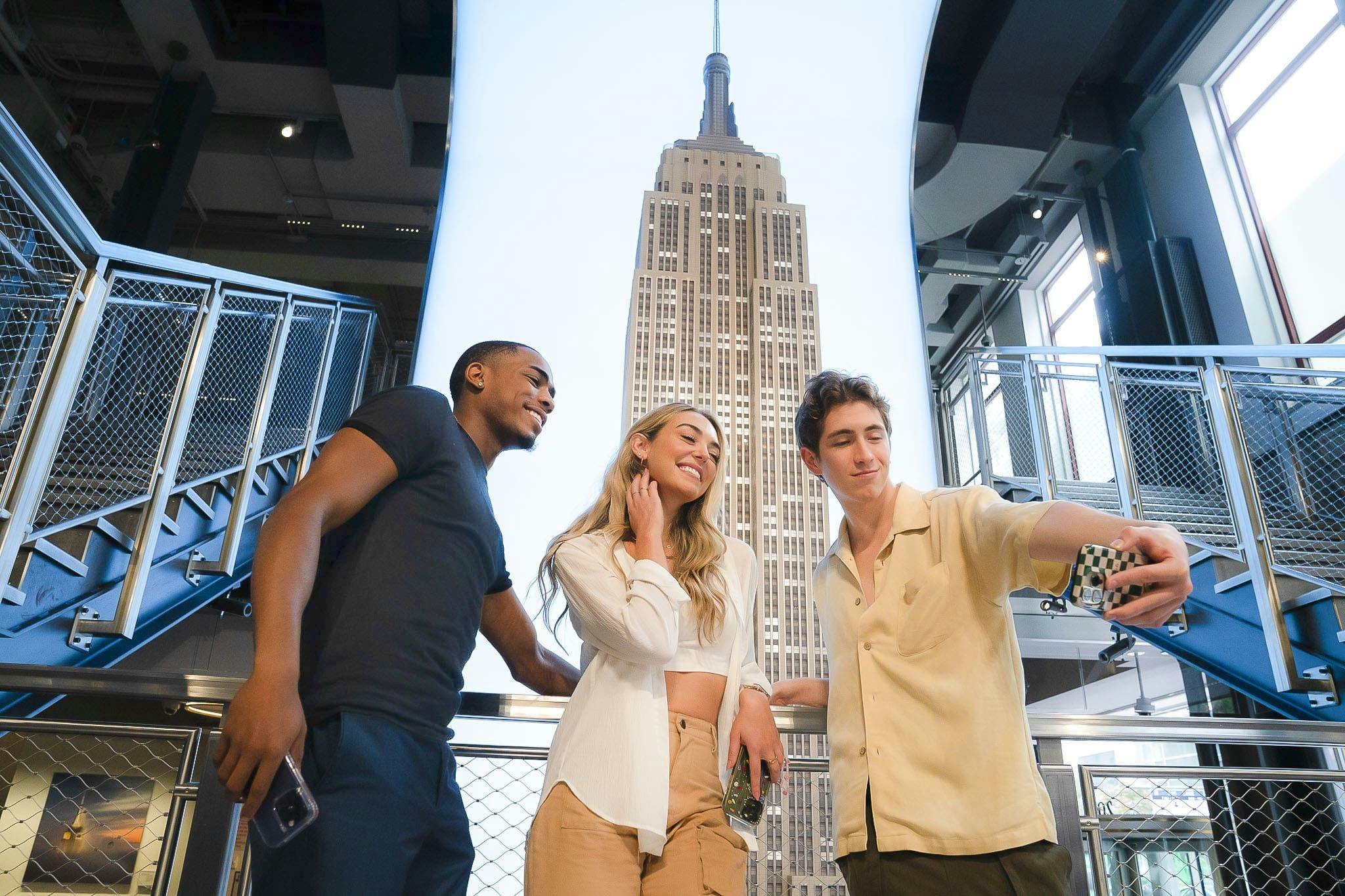 Three people posing for a selfie inside a building with a large display of the Empire State Building in the background.
