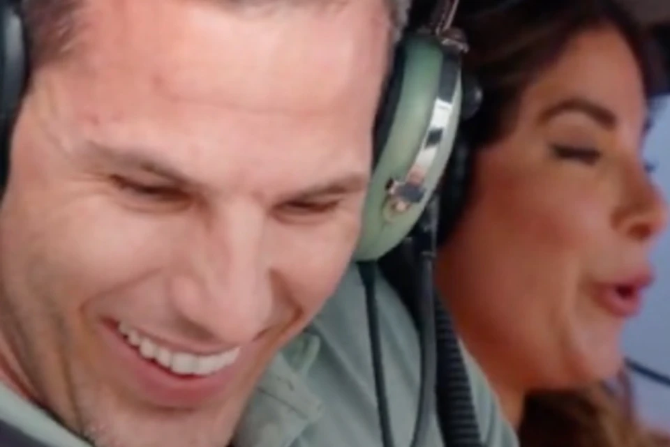 Two individuals wearing headsets sit in a cockpit, one smiling and the other speaking animatedly.