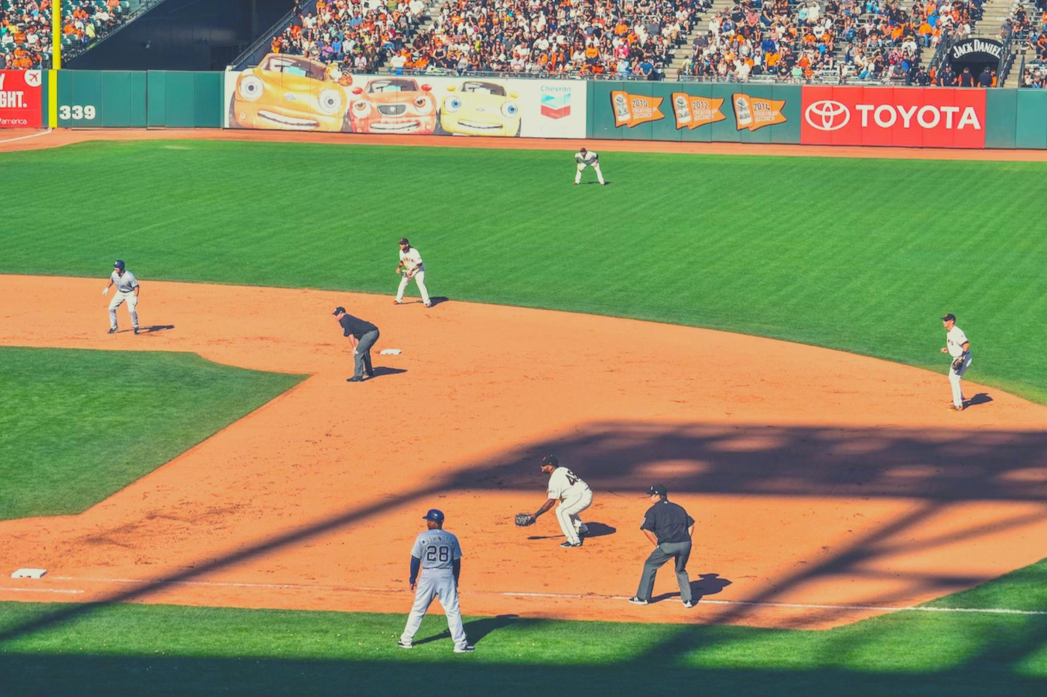Baseballkamp i gang med spillere på banen og en menneskemængde på tribunen. Reklamebannere er synlige i baggrunden.