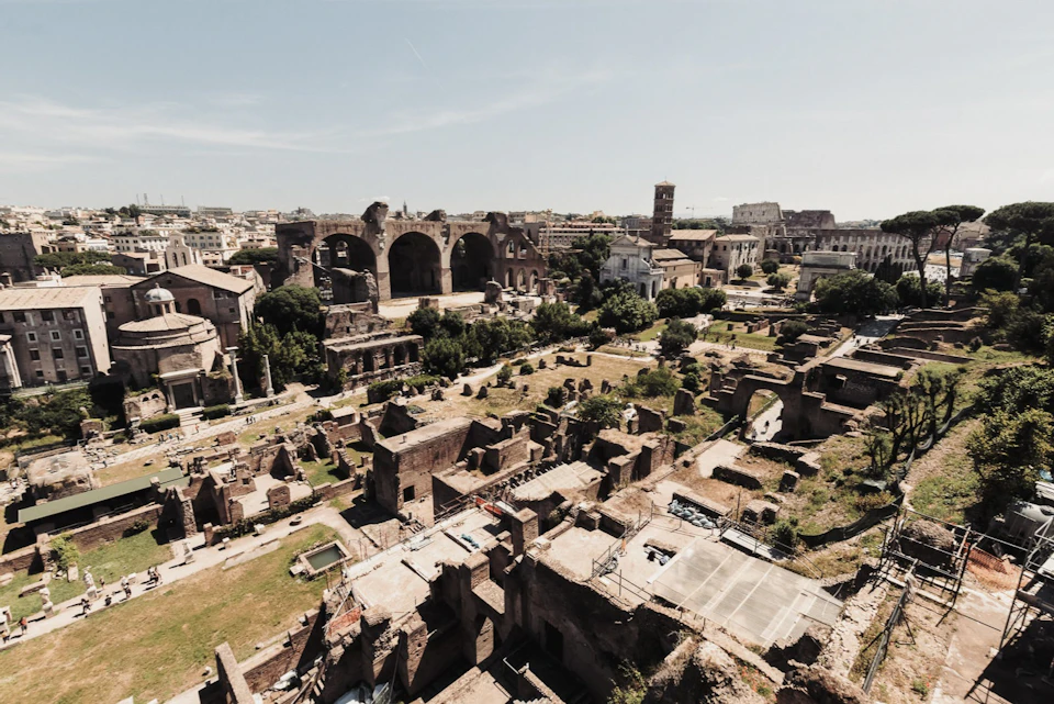 Aerial view of ancient Roman ruins with arches, scattered structures, and greenery under a clear sky.