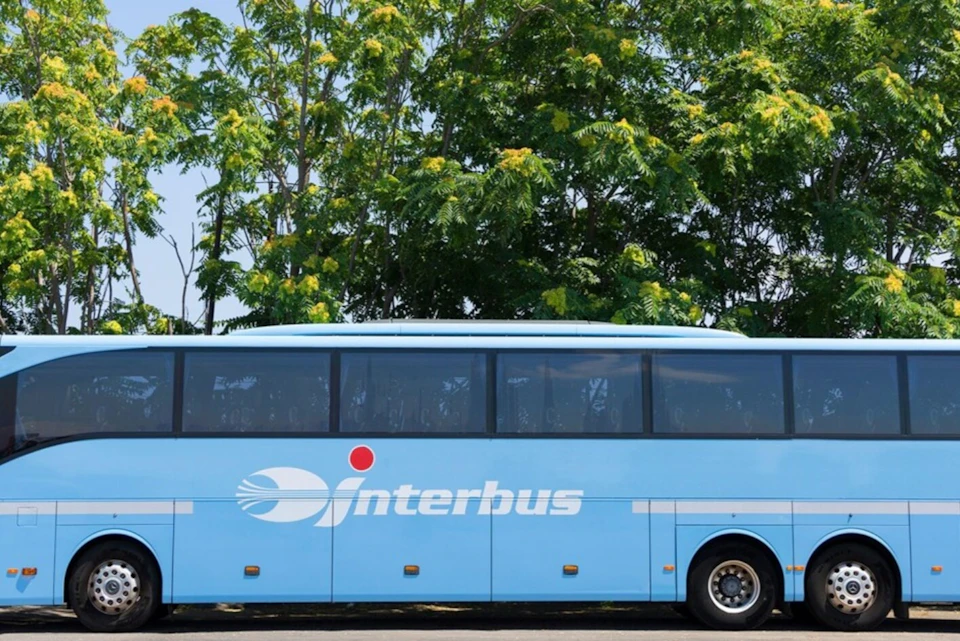 A blue bus with "Interbus" branding parked in front of lush green trees.