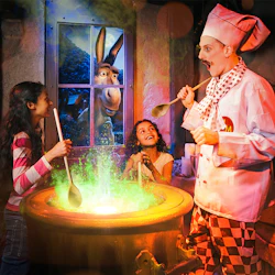 Två tjejer och en kock rör om i en glödande grön kittel, med en animerad åsna som tittar genom ett fönster bakom dem.