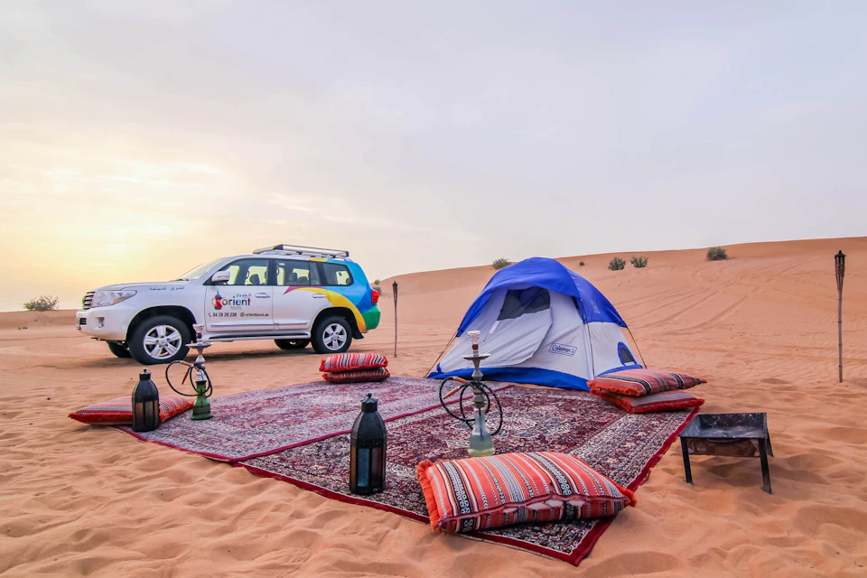 Desert campsite with a tent, colorful cushions on rugs, hookahs, lanterns, and an SUV parked nearby. Sparse vegetation in the background.
